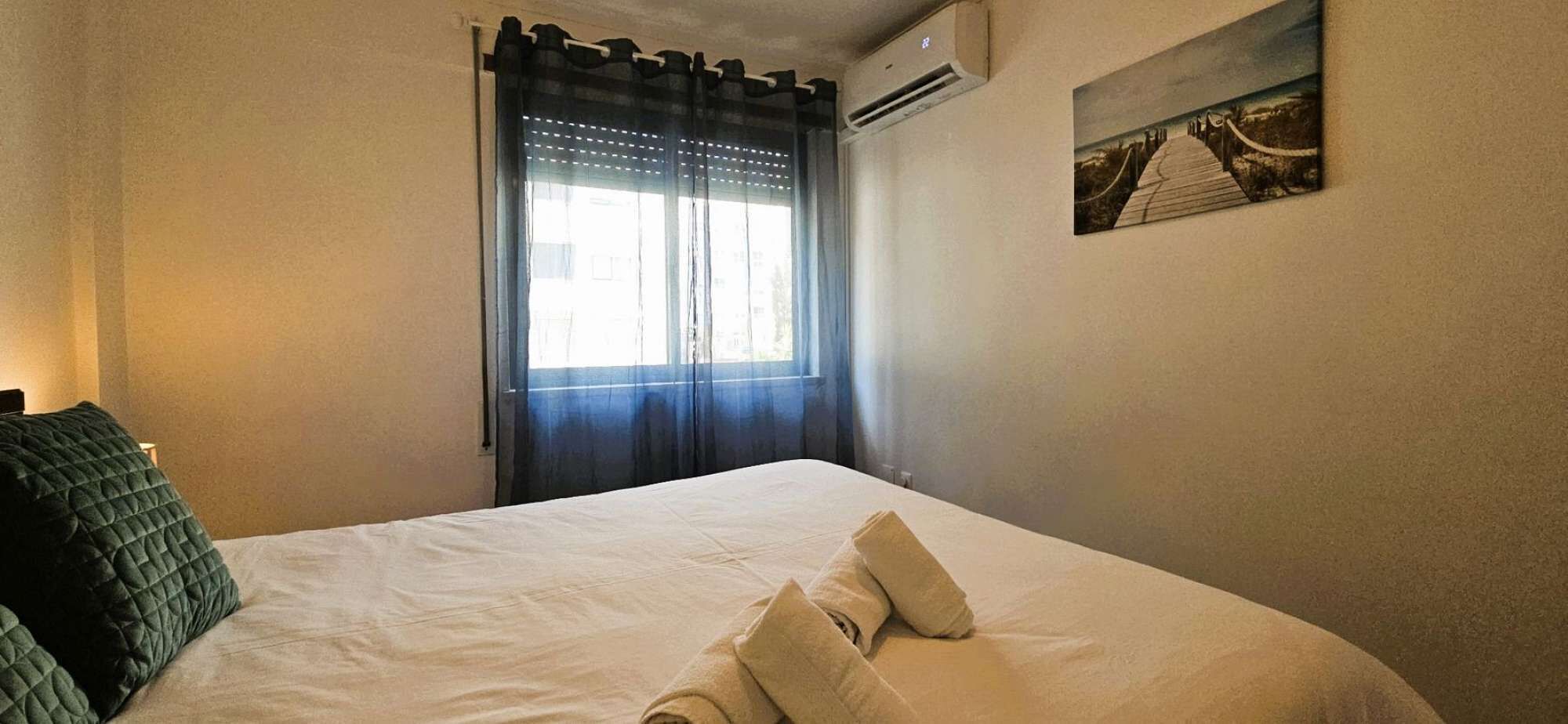 Un lit bien fait, avec des oreillers, une fenêtre avec des rideaux et un climatiseur fixé au mur.