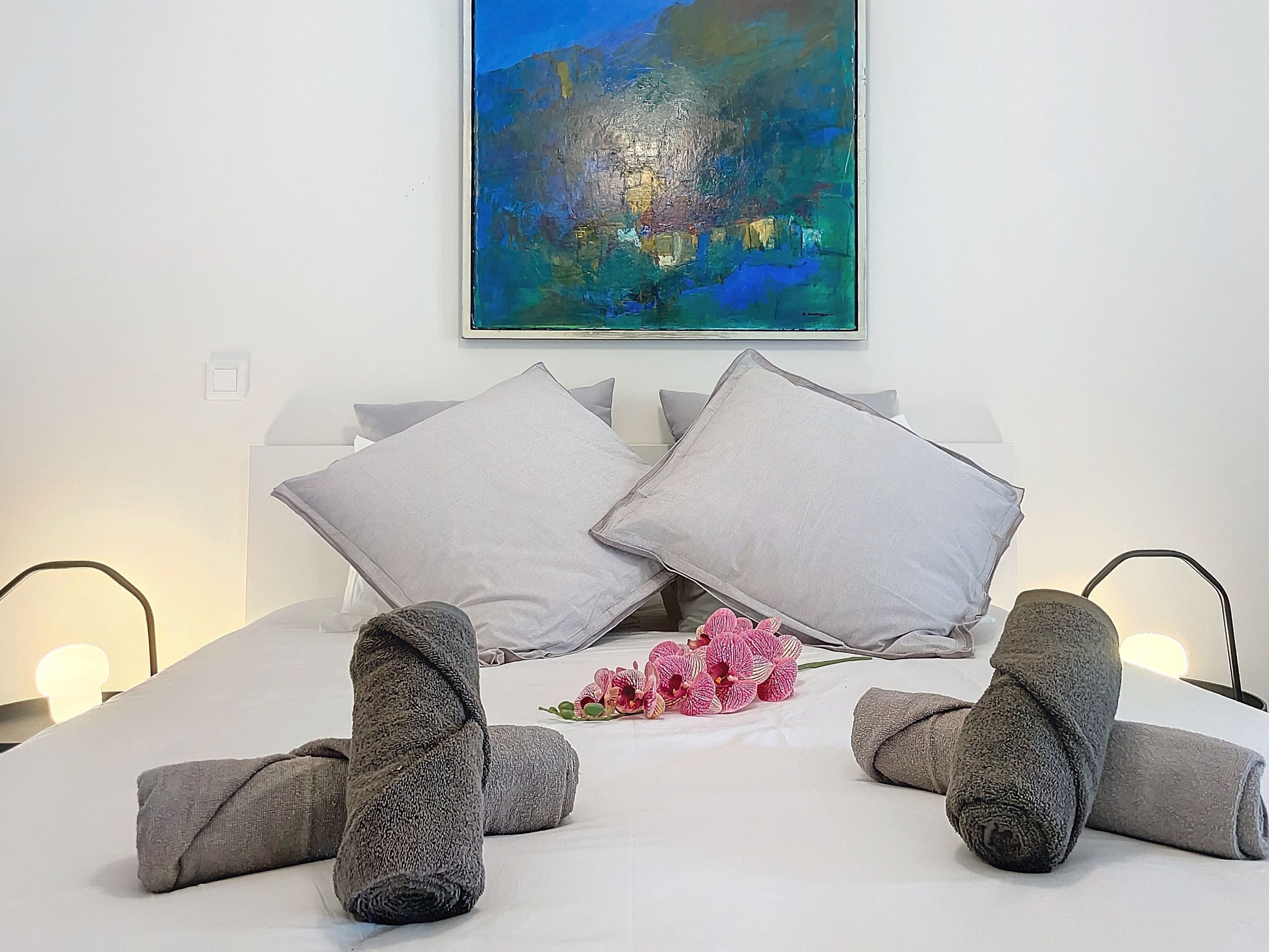 Een netjes opgemaakt bed met grijze kussens en opgerolde handdoeken, een schilderij van een stadsgezicht aan de muur, en een klein boeket roze bloemen.