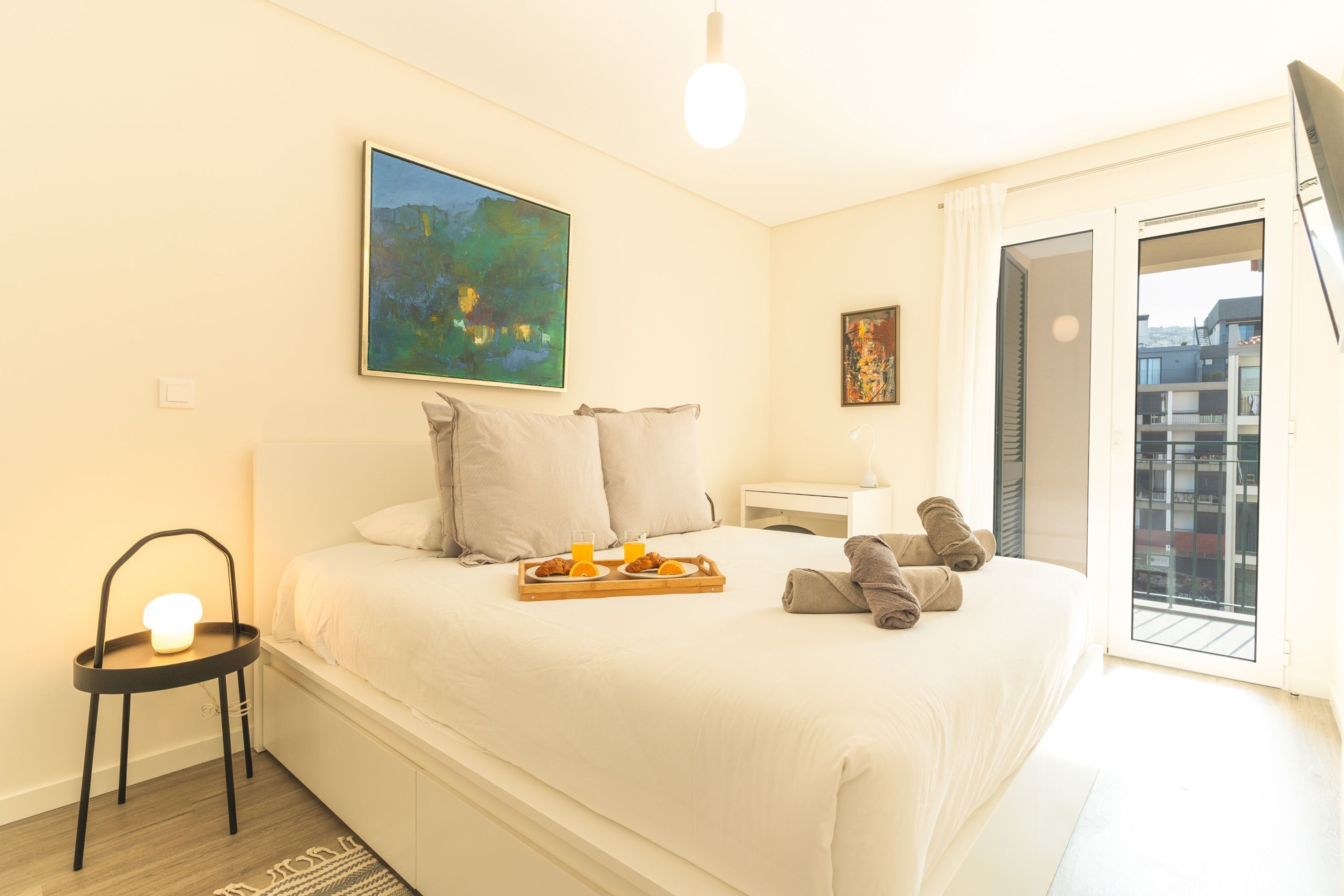 Habitación de hotel con una cama, una mesita de noche, una silla, una bandeja con desayuno y un cuadro en la pared.