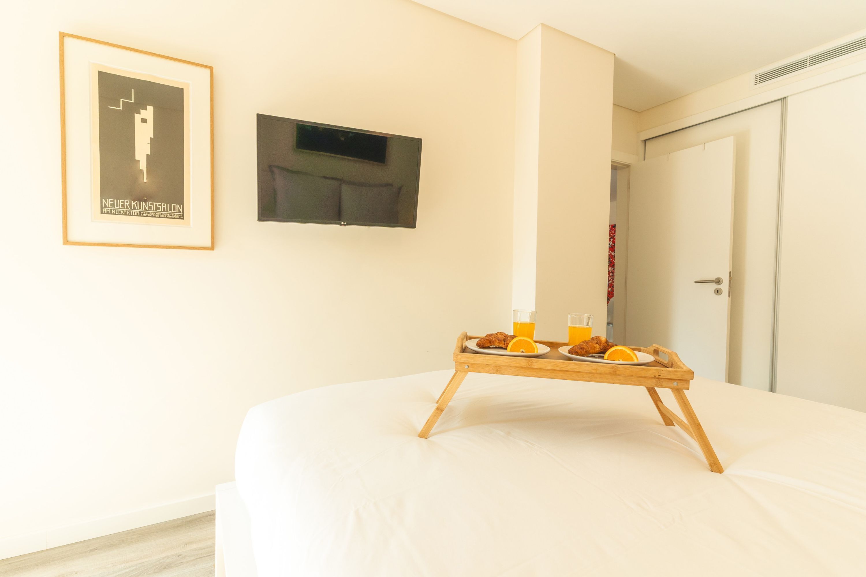 Habitació d'hotel amb un llit, un safata de croissants i suc d'aranja, un cartell emmarcat i una televisió penjada a la paret.