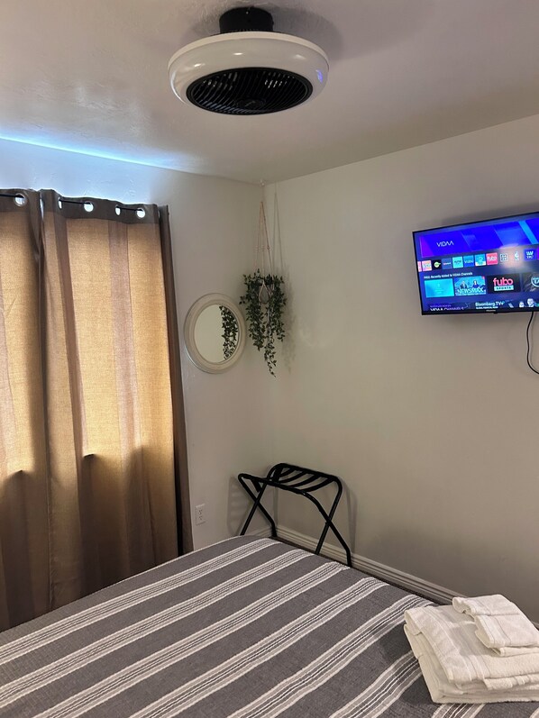 Une chambre d’hôtel avec un lit, un téléviseur fixé au mur, un ventilateur de plafond, une fenêtre avec des rideaux et une petite plante.