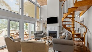 Ein geräumiges Wohnzimmer mit Kamin, einer Wendeltreppe aus Holz und großen Fenstern mit Blick ins Freie.