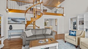 Ein geräumiges Wohnzimmer mit einer Holztreppe, einem grauen Sofa, einem beige farbenen Sessel und einem Couchtisch.