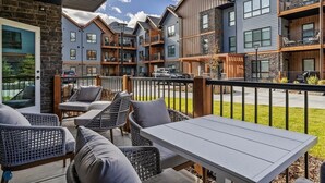 Un área de estar al aire libre moderna con mobiliario de mimbre y una mesa, con vistas a una hilera de edificios de varios pisos con balcones.