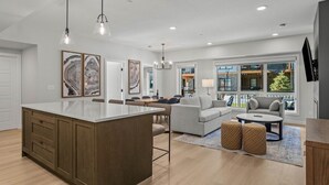 Una cocina moderna con una isla central, comedor con sillas, un sofá y una mesa de centro. Hay lámparas colgantes, un ventanal con vistas y un televisor de pantalla plana montado en la pared.