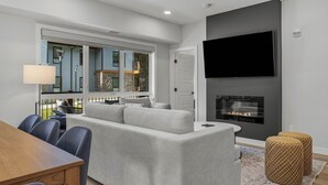 Una sala de estar moderna con un sofá seccional gris, una mesa de comedor de madera y sillas. Hay una chimenea, un televisor de pantalla plana montado en la pared y un ventanal con vistas a un edificio azul.