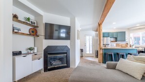 A modern living room with a fireplace, a television mounted on the wall, and a kitchen area with blue cabinets and a bar. - Cute modern 2 bed retreat, a perfect basecamp for your adventures! (Fraser)