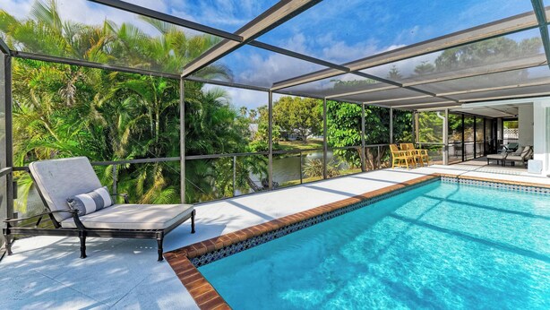 A pool area with a glass-enclosed deck, a lounge chair, and a view of lush greenery and a body of water.
