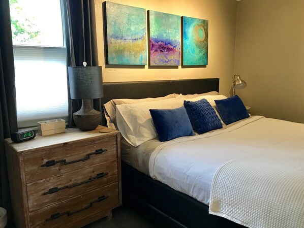 A hotel room with a bed, bedside lamp, clock, wooden nightstand, and abstract wall art.