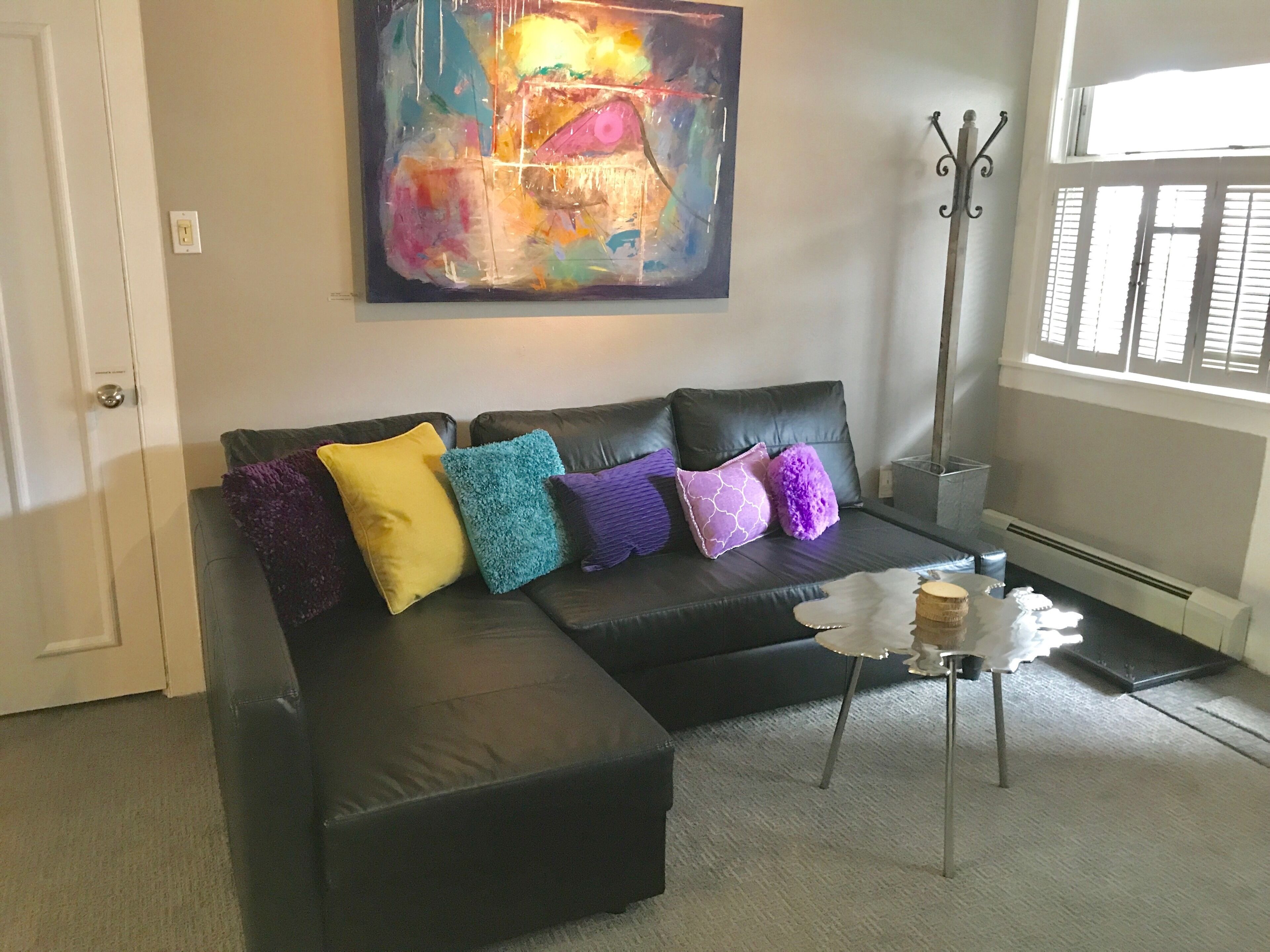 Sala de estar com sofá de couro preto, almofadas coloridas, mesa de centro branca e uma grande pintura abstrata na parede.
