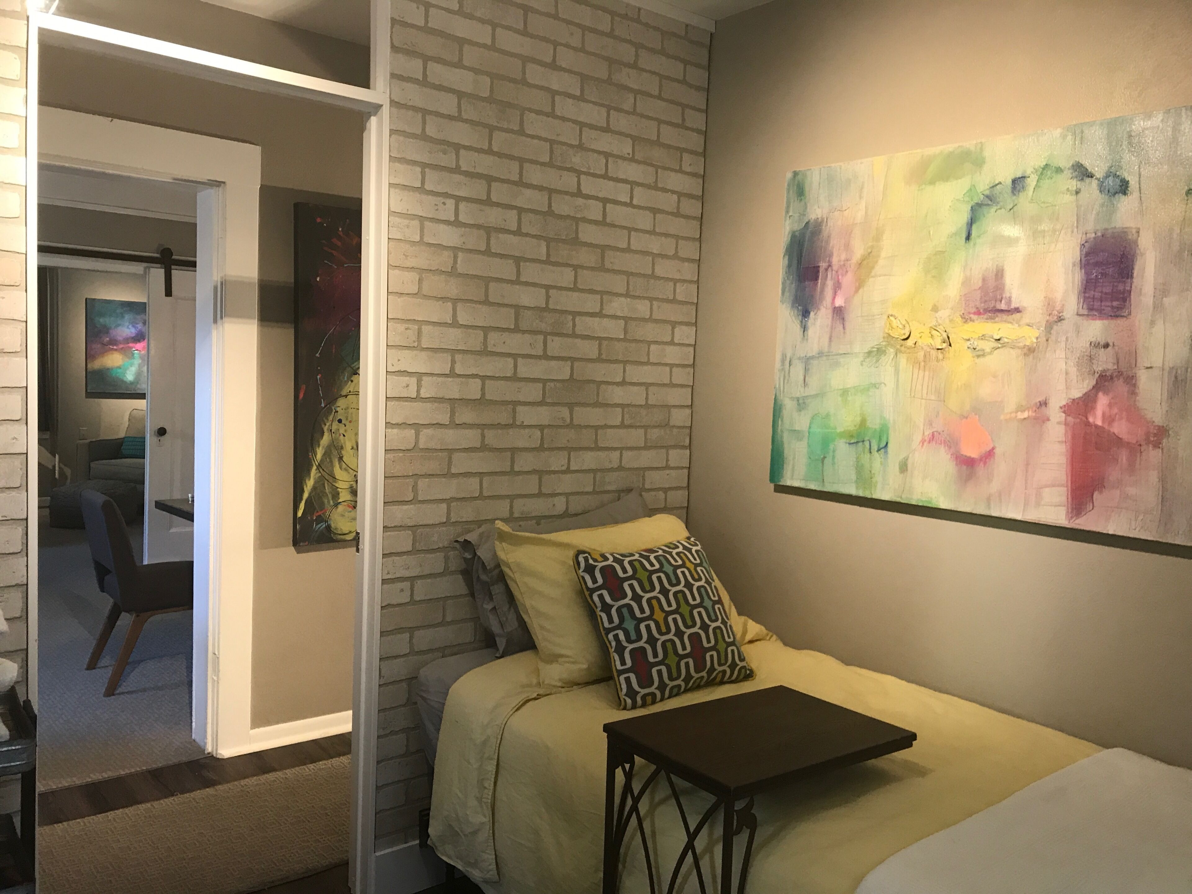 Een slaapkamer met een metselwerkmuur, een bed met gekleurde kussens, een bijzettafeltje en een kleurrijk abstract schilderij.