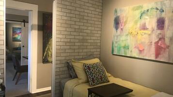 Een slaapkamer met een metselwerkmuur, een bed met gekleurde kussens, een bijzettafeltje en een kleurrijk abstract schilderij.