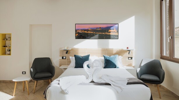 A hotel room with a bed, two chairs, a TV on the wall, and a view of a cityscape.