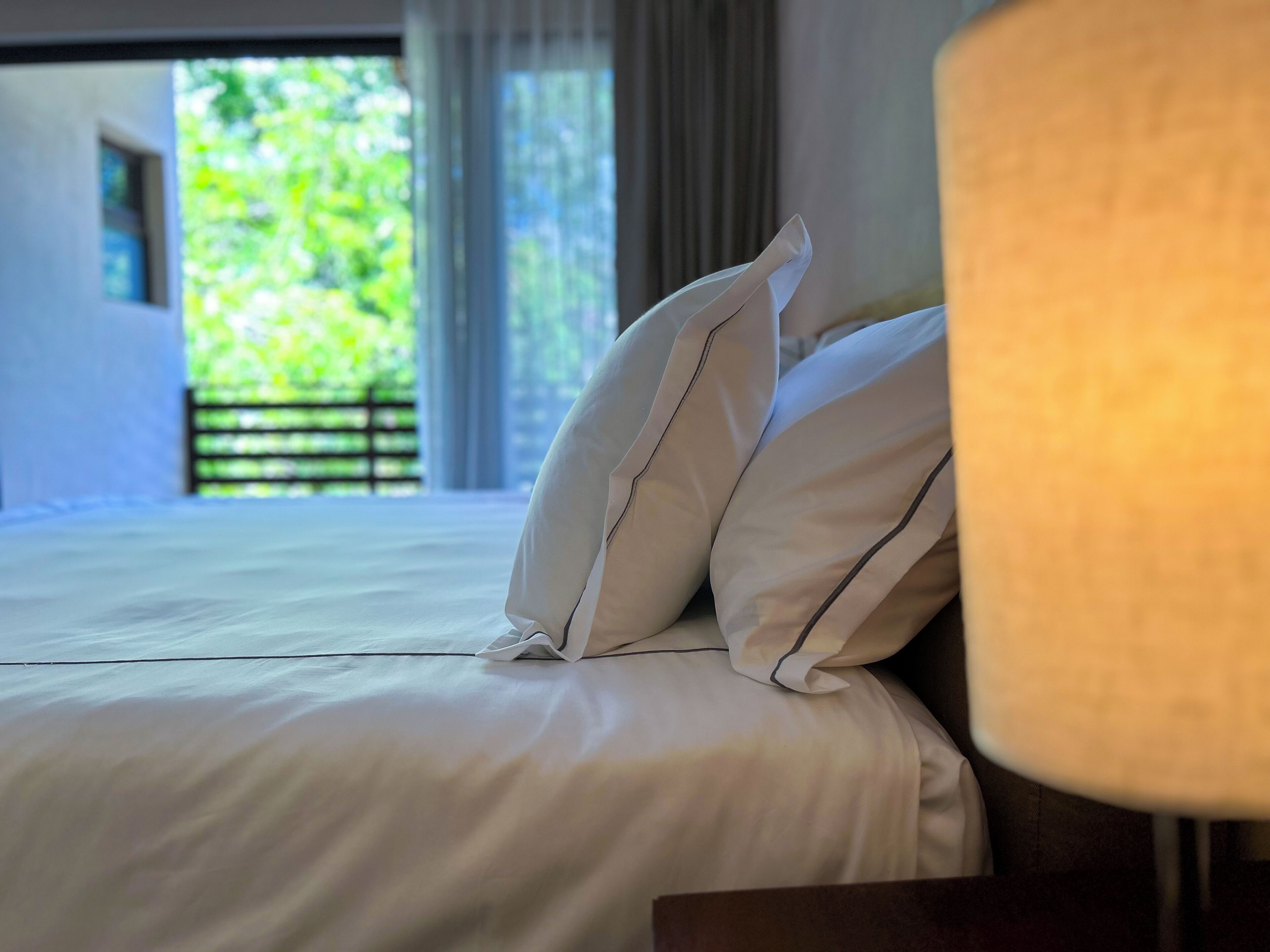 A bed with white bedding and pillows, a nightstand with a lamp, and a view of greenery outside.