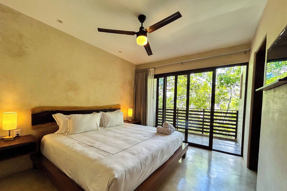 A spacious bedroom with a large bed, a ceiling fan, and a view of greenery through the window.