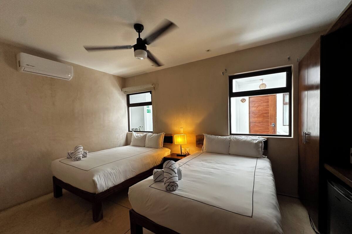 A hotel room with two beds, a ceiling fan, a window with a view, and a lamp.