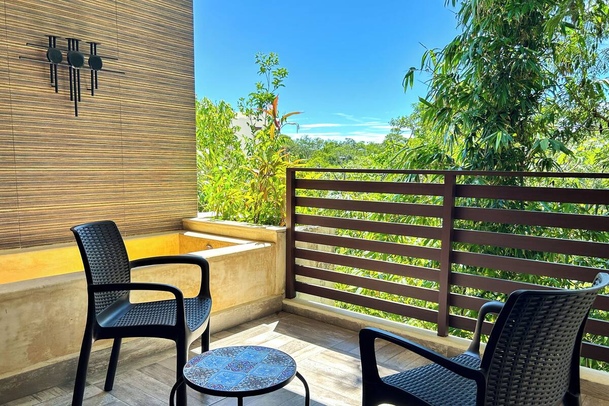 A balcony with two chairs and a small table, a wooden fence, and a view of greenery.