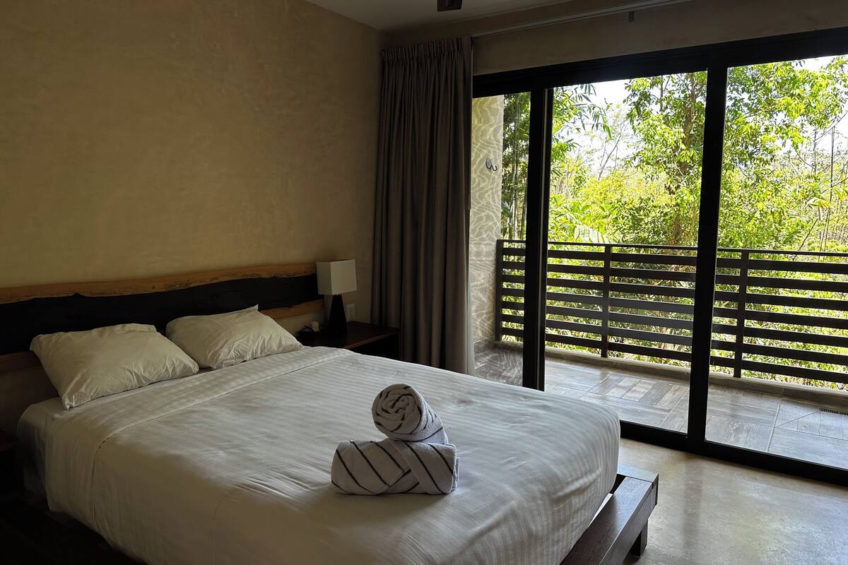 A hotel room with a large bed, a nightstand, a lamp, and a view of greenery through a balcony.