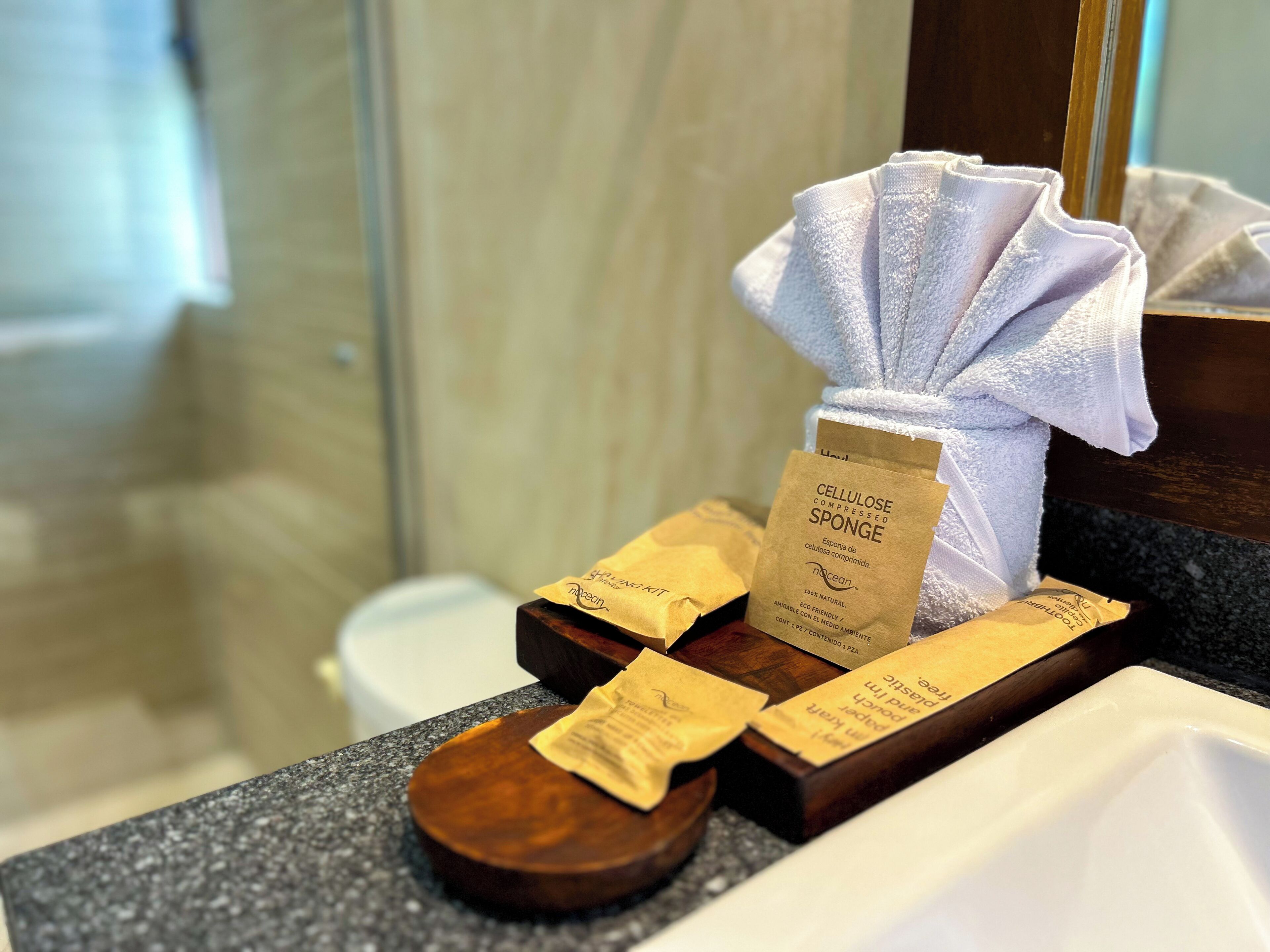 Ein Badezimmer mit einem weißen Waschbecken, einem Spiegel und einem Handtuchspender mit einem weißen Handtuch. Auf der Ablage liegen außerdem zwei braune Papiertüten.
