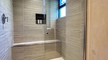 Un baño moderno con ducha de vidrio, un banco y un estante pequeño con artículos de aseo.