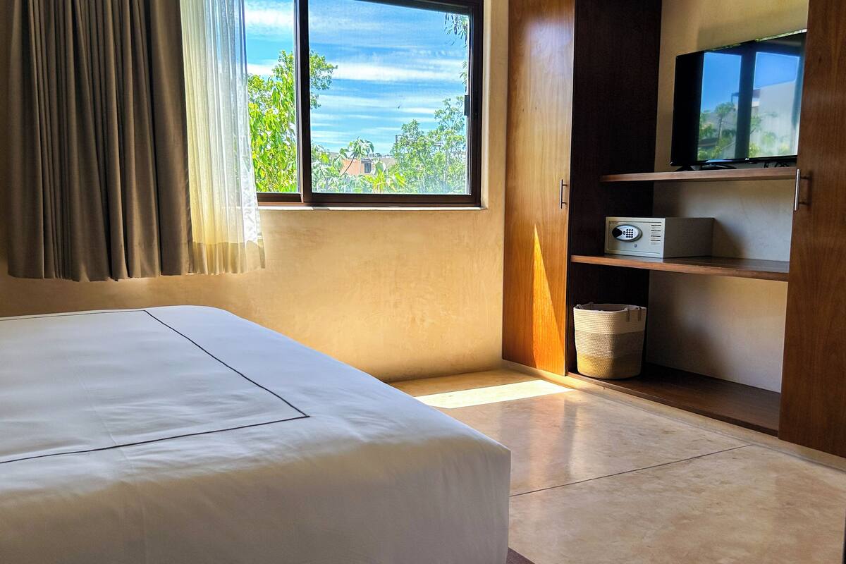 A hotel room with a bed, a window with curtains, a television on a wooden shelf, and a small radio.