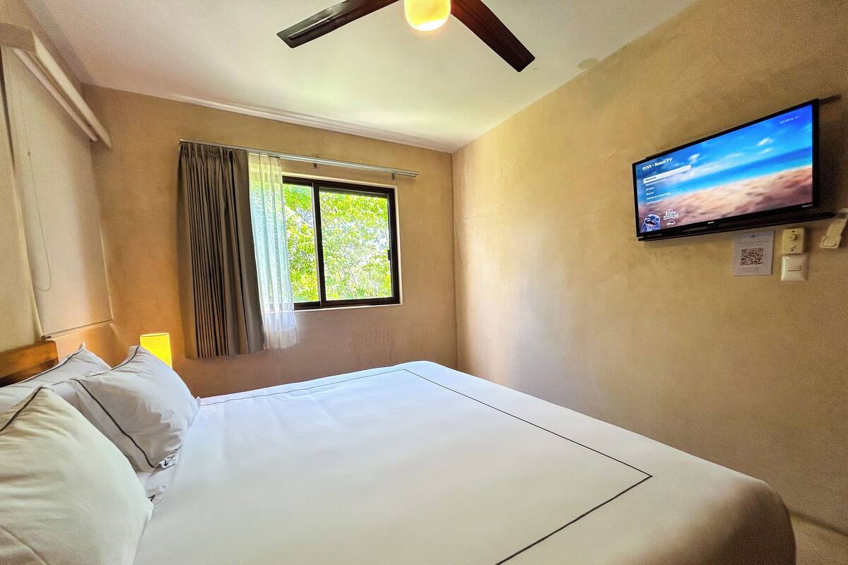 A hotel room with a bed, a window with curtains, a television on the wall, and a ceiling fan.