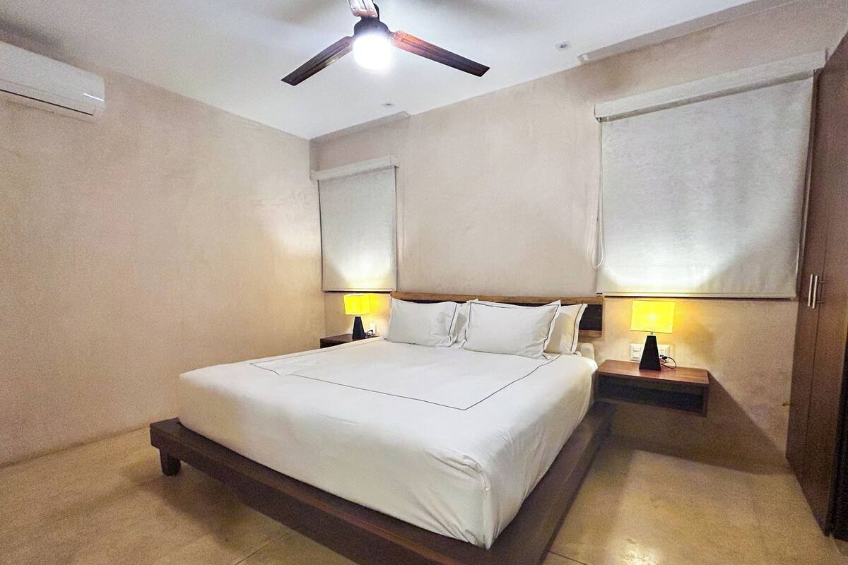 A bedroom with a large bed, two bedside tables with lamps, a ceiling fan, and a window with blinds.