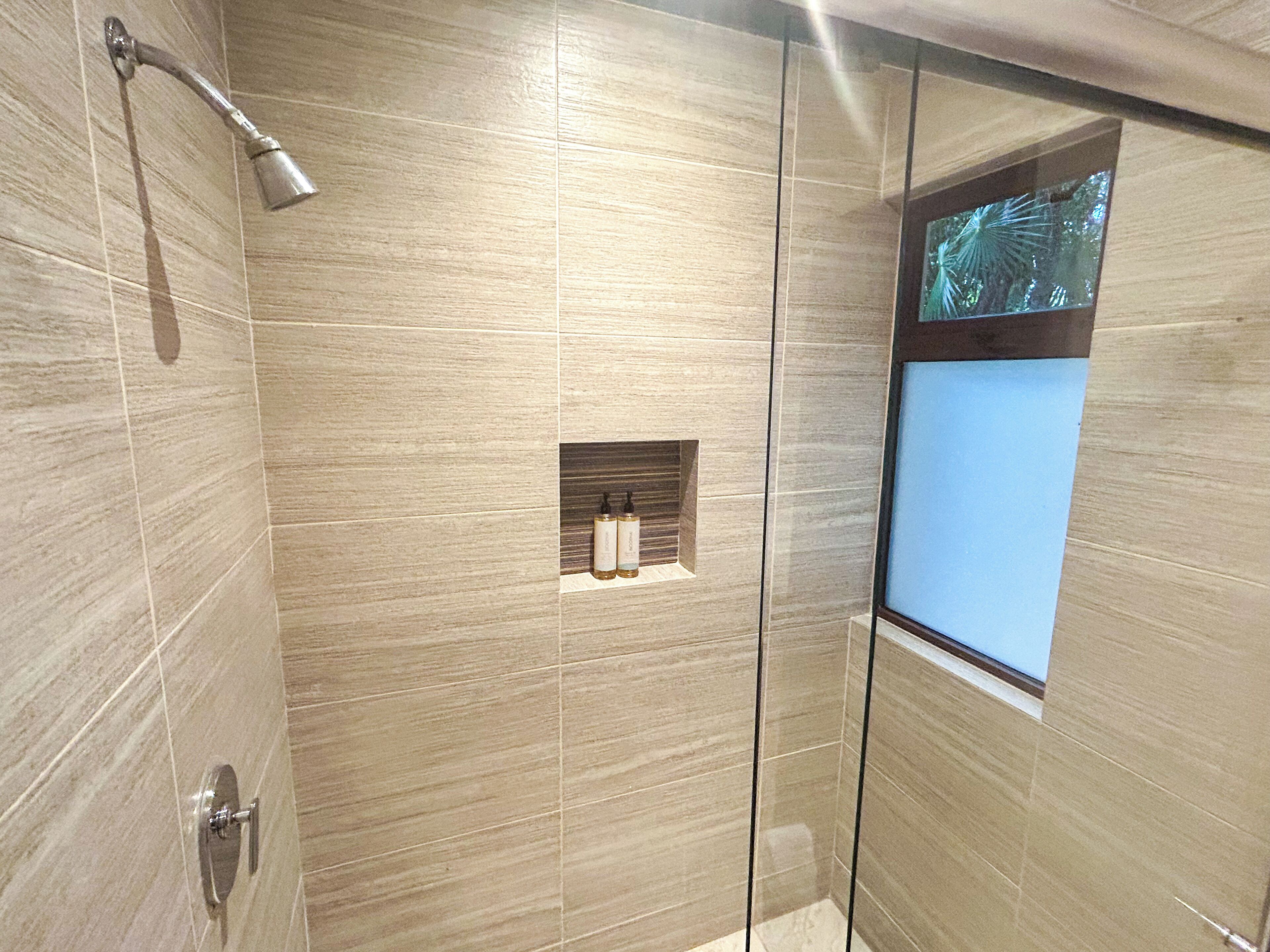 Une douche moderne avec une porte en verre, une étagère intégrée pour les articles de toilette et une fenêtre donnant sur des palmiers.