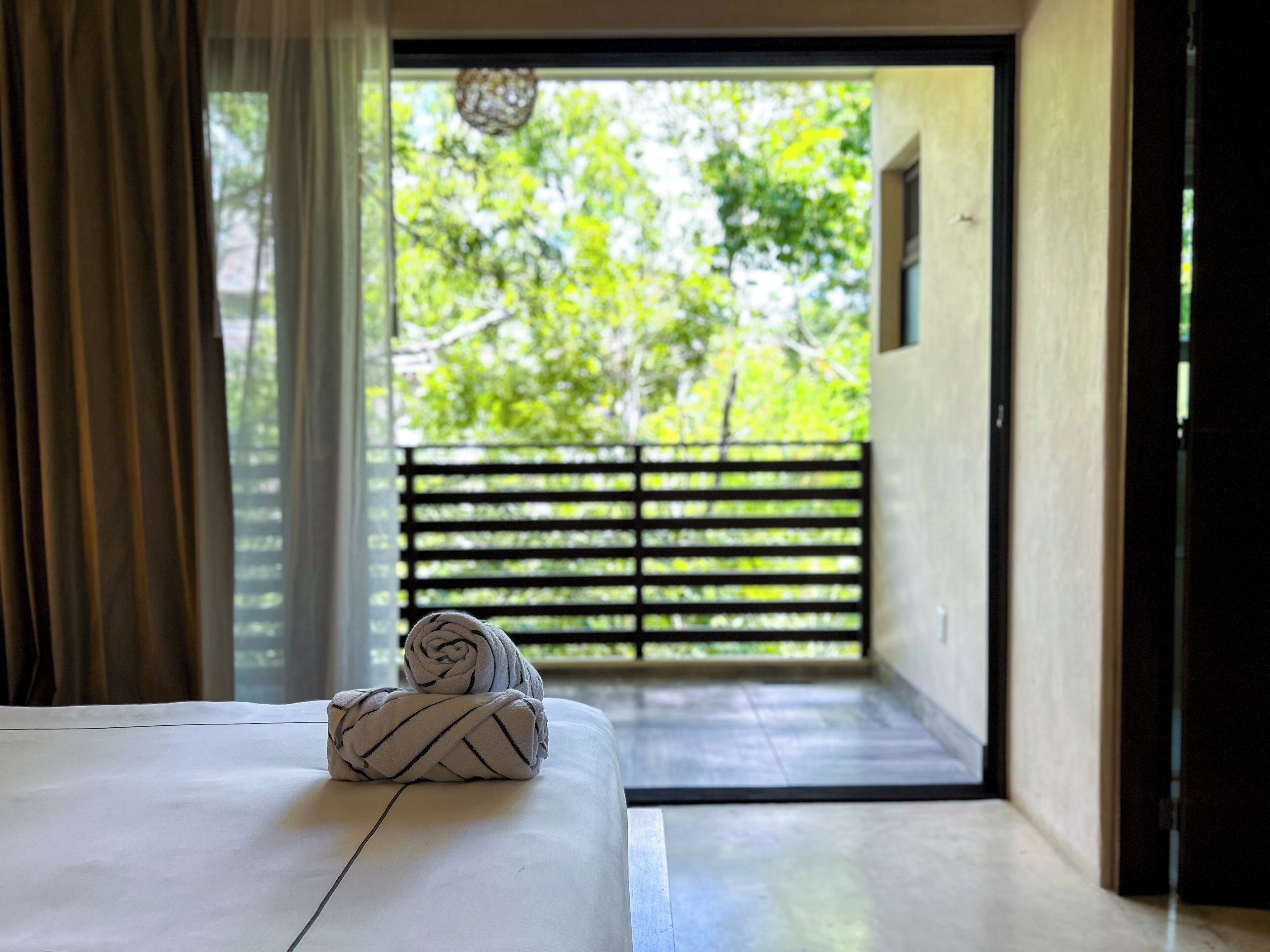 A room with a bed, a pair of towels, a view of greenery through a balcony door, and a hanging light fixture.