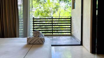 A room with a bed, a pair of towels, a view of greenery through a balcony door, and a hanging light fixture.