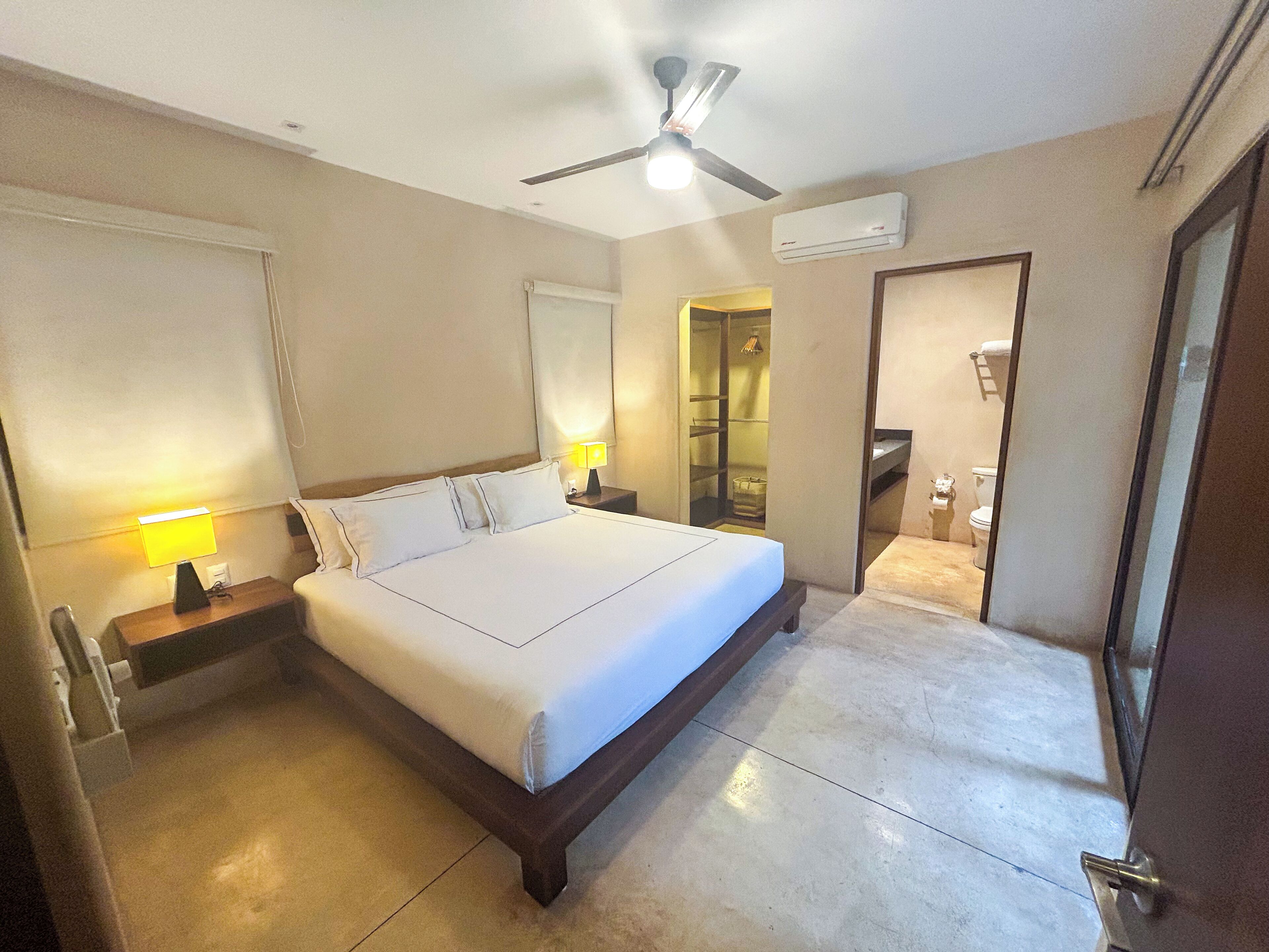 A hotel room with a large bed, a ceiling fan, a bedside lamp, and an air conditioning unit.