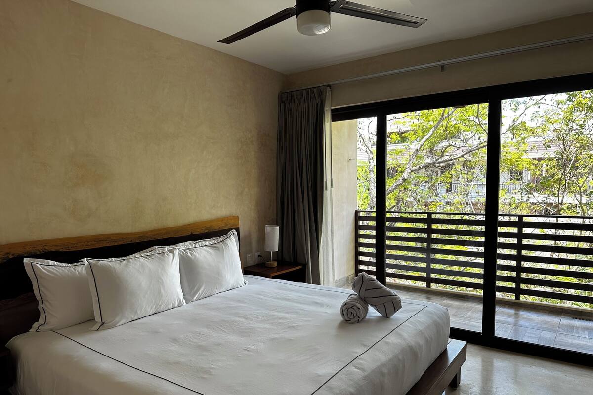 A hotel room with a bed, a ceiling fan, a nightstand, a lamp, and a view of greenery through large windows.