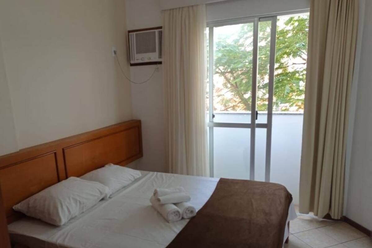 A hotel room with a bed, a wooden headboard, a window with curtains, and a view of greenery outside.