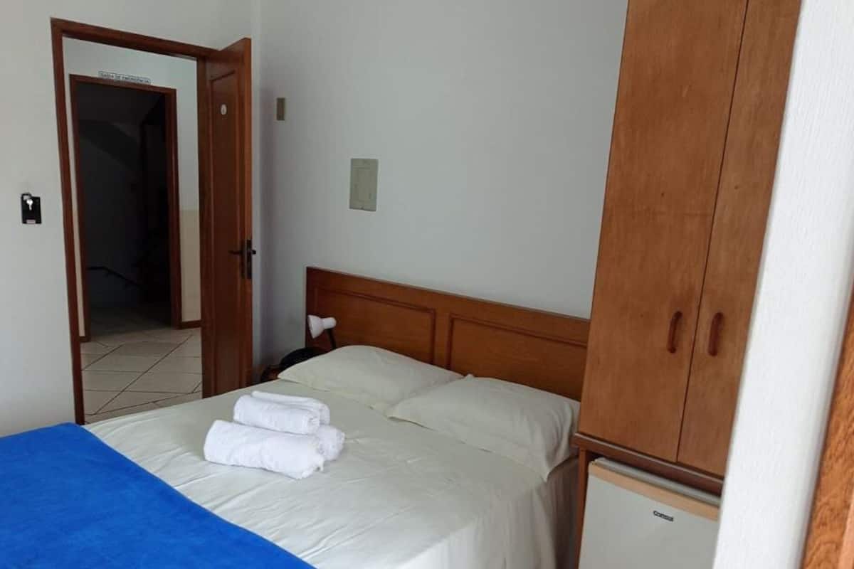A hotel room with a bed, a wardrobe, a small refrigerator, and a door leading to another room.
