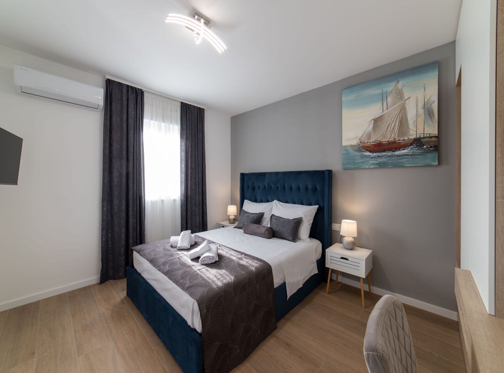 A modern hotel room with a large bed, two bedside tables with lamps, a painting of a sailboat, and a window with curtains.