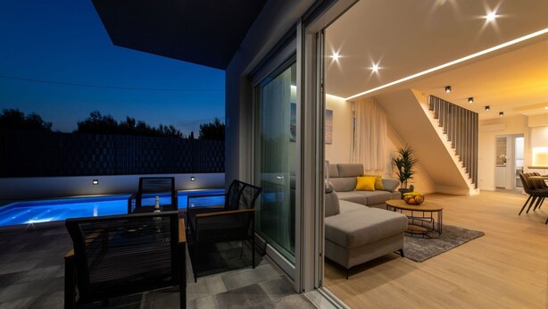 A modern living room with a glass door leading to a pool area, a staircase, and a dining area with a table and chairs.