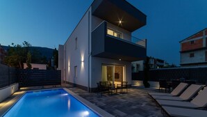 A modern two-story house with a swimming pool, outdoor seating, and a well-lit balcony.
