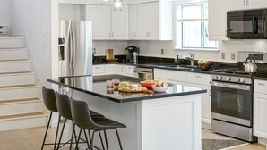 Una cocina moderna con armarios blancos, electrodomésticos de acero inoxidable, una isla central con taburetes y un amplio ventanal que proporciona luz natural.