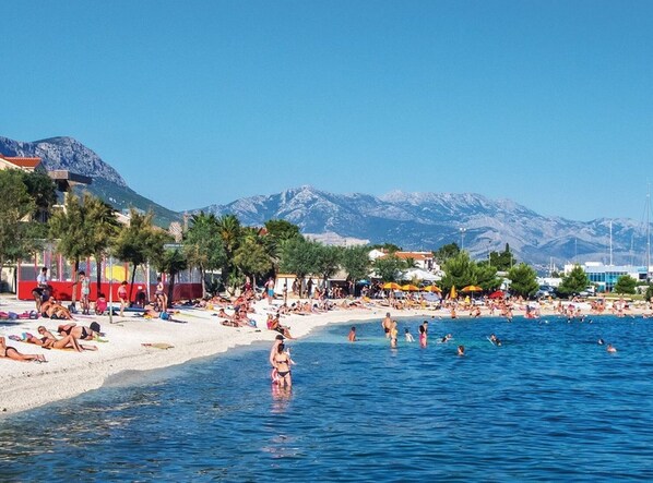 Relax on the beach with palm trees and umbrellas providing shade, surrounded by calm waters and scenic mountains.