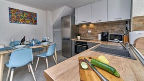 Een moderne keuken met een houten aanrecht, een spoelbak en een koelkast. Er staan twee blauwe stoelen bij een eettafel met wijnglazen en flessen.