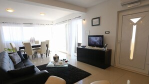 A modern living room with a black sofa, a glass coffee table, and a TV on a black stand. There is a dining area with a table and chairs in the background.