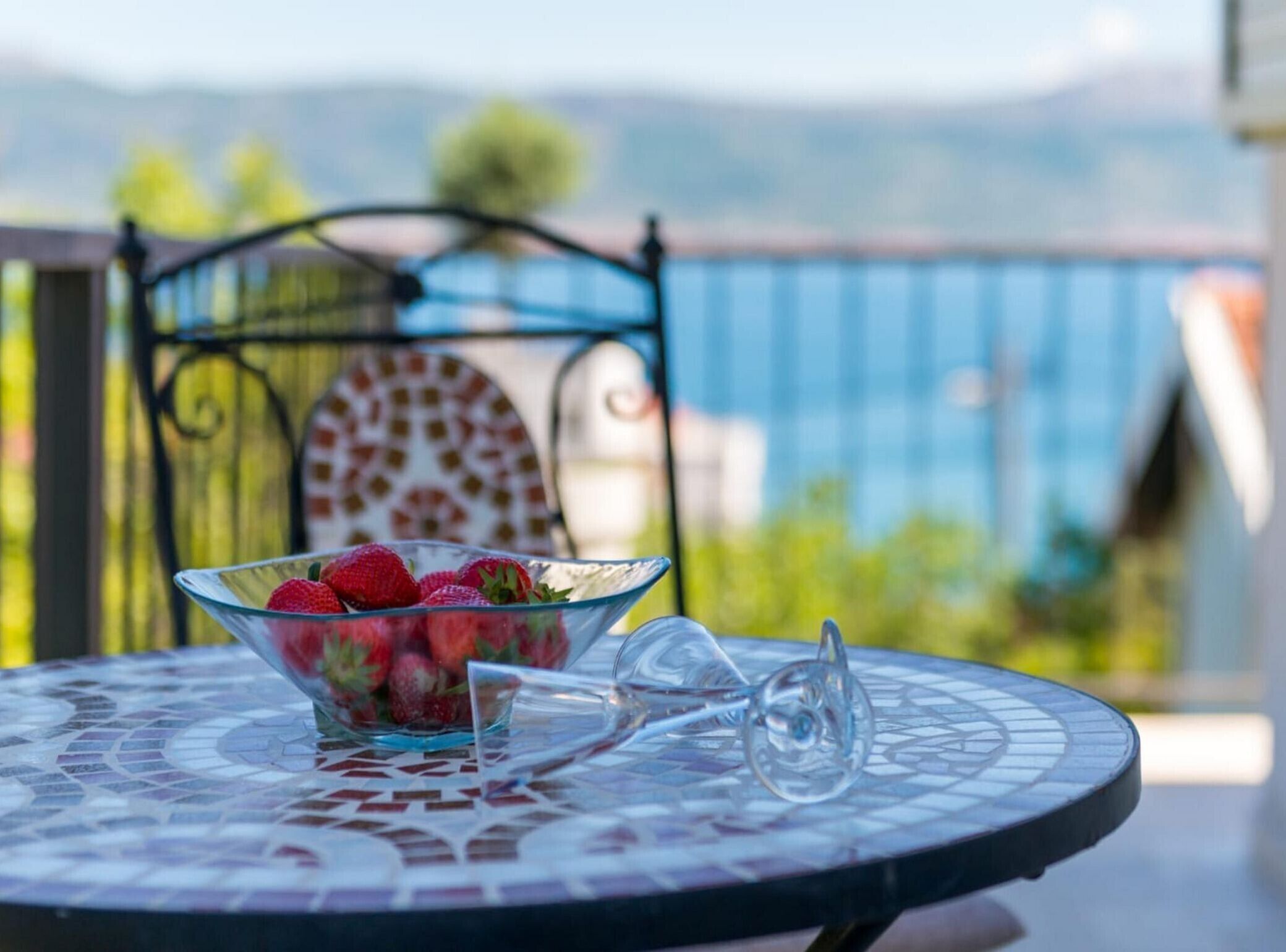 Un saladier en verre rempli de fraises est posé sur une table à motifs de mosaïque, avec un verre à vin à côté. L’ensemble est placé sur un balcon d’où l’on peut admirer une vue sur les montagnes et une rambarde bleue.