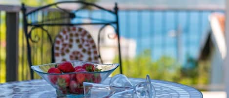 A glass bowl of strawberries on a mosaic table with a wine glass beside it, set on a balcony with a view of mountains and a blue railing.