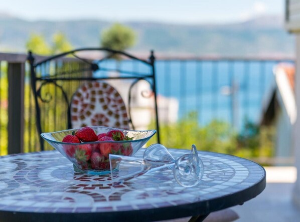 A glass bowl of strawberries on a mosaic table with a wine glass beside it, set on a balcony with a view of mountains and a blue railing.