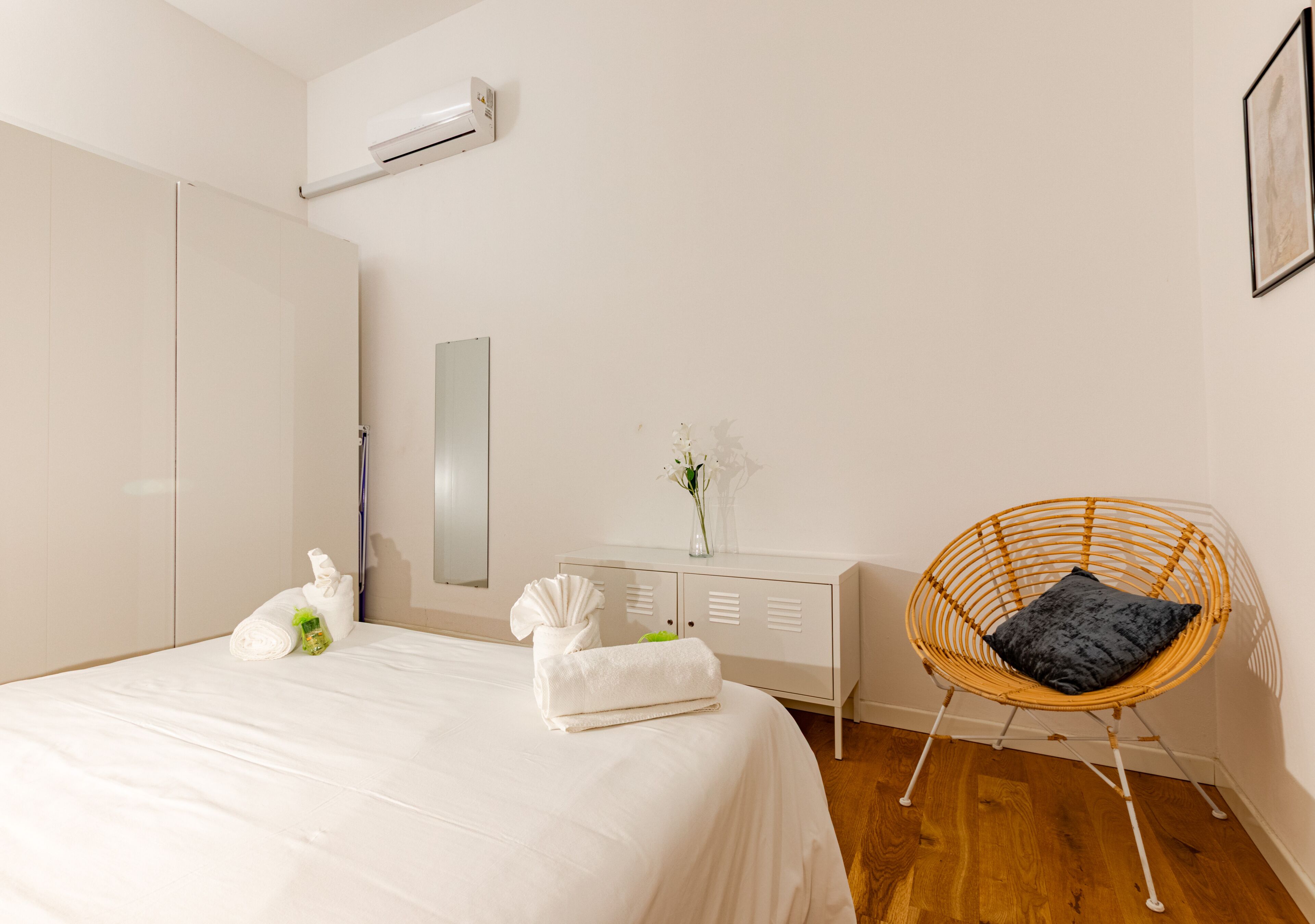 A bedroom with a bed, a white wardrobe, a wicker chair with a cushion, a dresser, and a mirror.