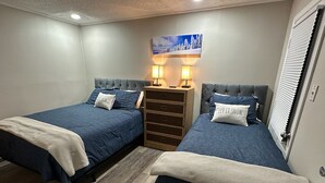 Un dormitorio con dos camas, una mesita de noche, una lámpara de pared y una ventana con persianas.