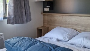 Una cama bien hecha con colcha y almohadas azules, una mesita de noche con lámpara y una ventana con cortinas.