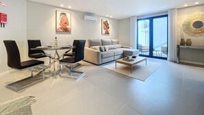 Un soggiorno moderno con zona pranzo, divano e tavolino. Due quadri alle pareti, una lampada a soffitto e vista all'esterno attraverso un'ampia finestra.