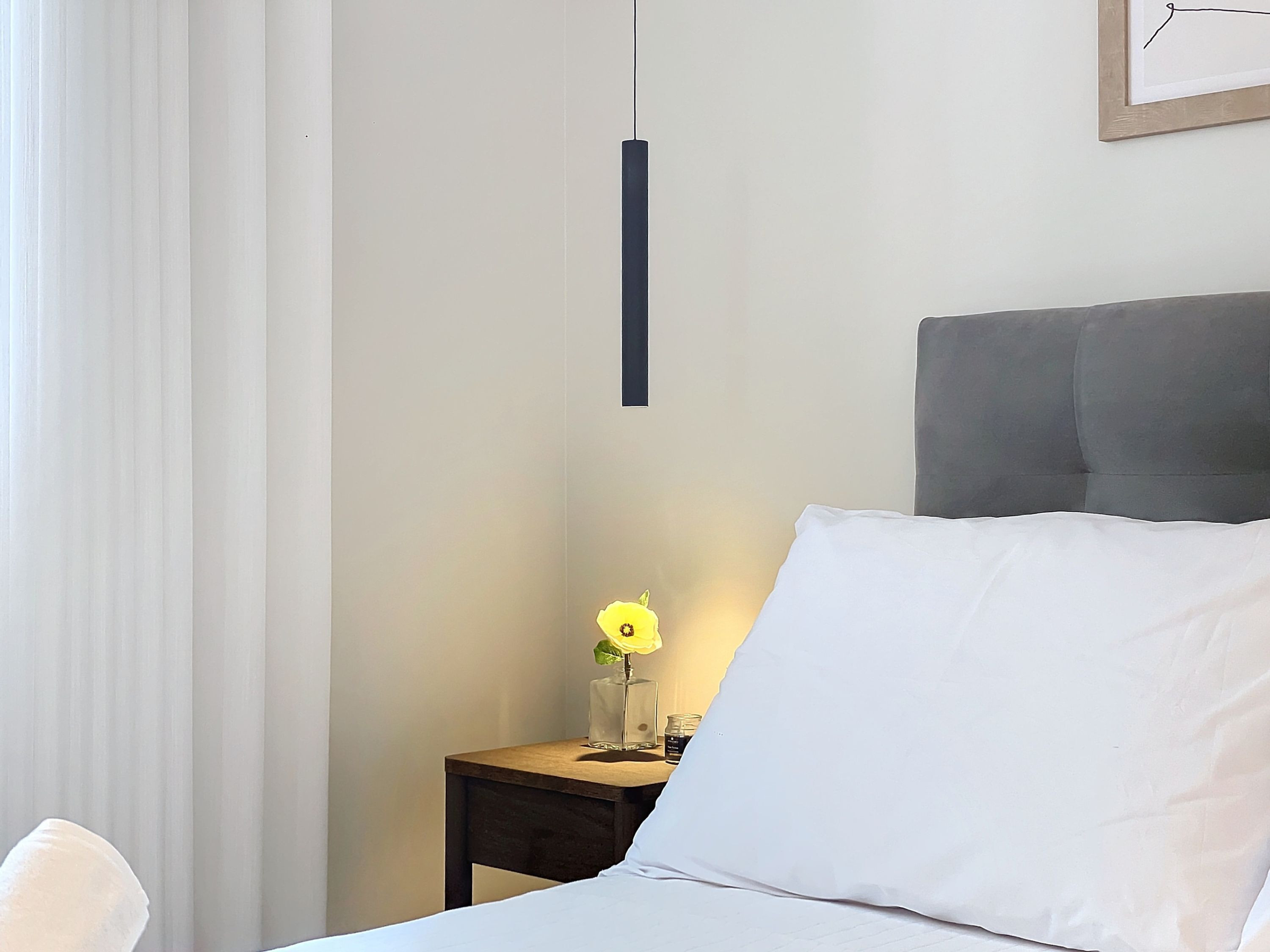 A neatly made bed with white linens, a bedside table with a vase of yellow flowers, and a hanging pendant light.