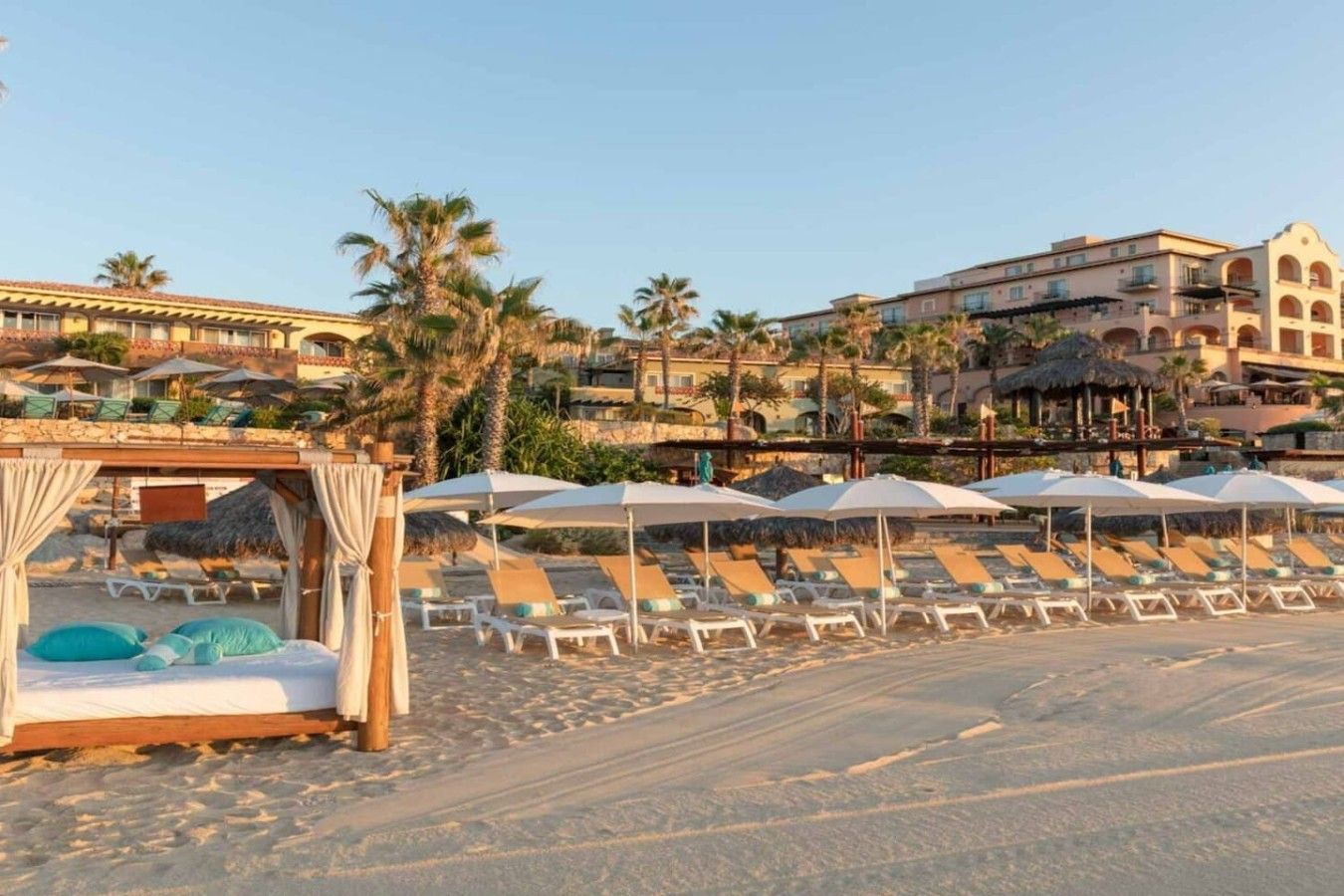 A beachside hotel with sun loungers and umbrellas, a cabana, and a building with arches in the background.